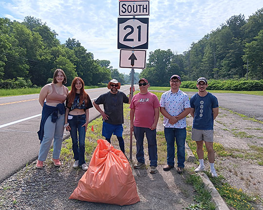 group of people standing next to a road sign with a bag of trash in front of them