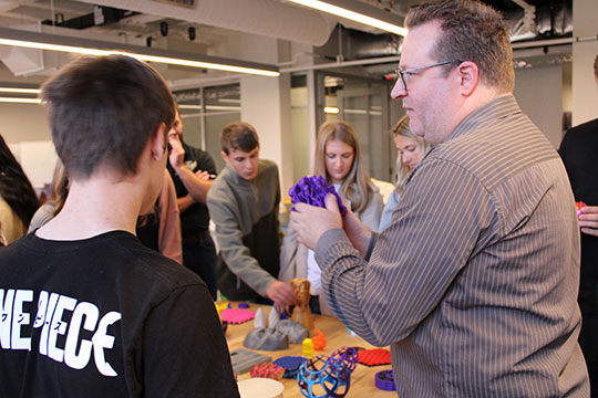 a man holding an object and talking to a group of people gathered around a table