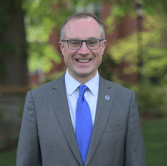 headshot of man in suit and tie, wearing glasses, smiling