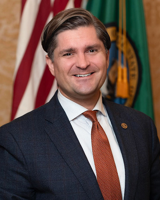 headshot of man wearing suit and tie smiling, in front of an American flag