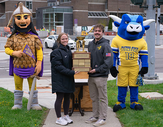a young man and young woman holding a trophy, with two mascots standing next to them