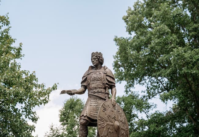 A bronze statue of a historical figure wearing armor, holding a stylized shield, and gesturing with one hand. Surrounded by lush green trees under a blue sky.