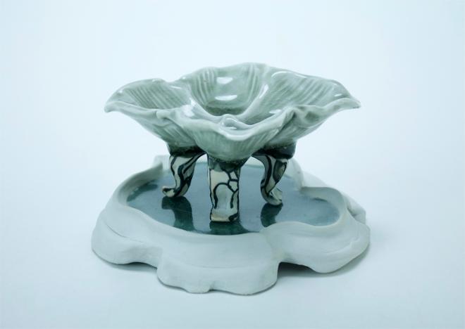 A small four legged dish atop a celadon pool in a porcelain trivet. 