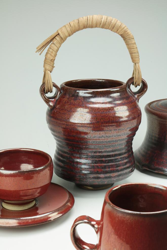 A group of iron red pots containing a cup and saucer, mug, and a tall vase-like form with a tan woven reed handle.    