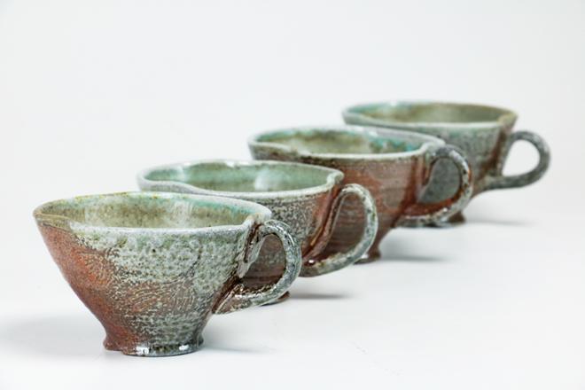 A line of four soda fired teacups with a carved ginkgo leaf design.  The surface shows orange and brown atmospheric flashing with deposits of soda glaze, the interiors are glazed in a green celadon.  