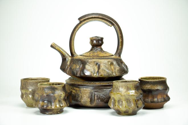 A wood-fired teapot sits on a stand, surrounded by a set of four matching cups.  The surfaces are whittled in appearance and color, as if they were carved from wood instead of thrown on the potter’s wheel.  Jade green and gold natural ash glaze cover the dark brown surface of the clay body.   