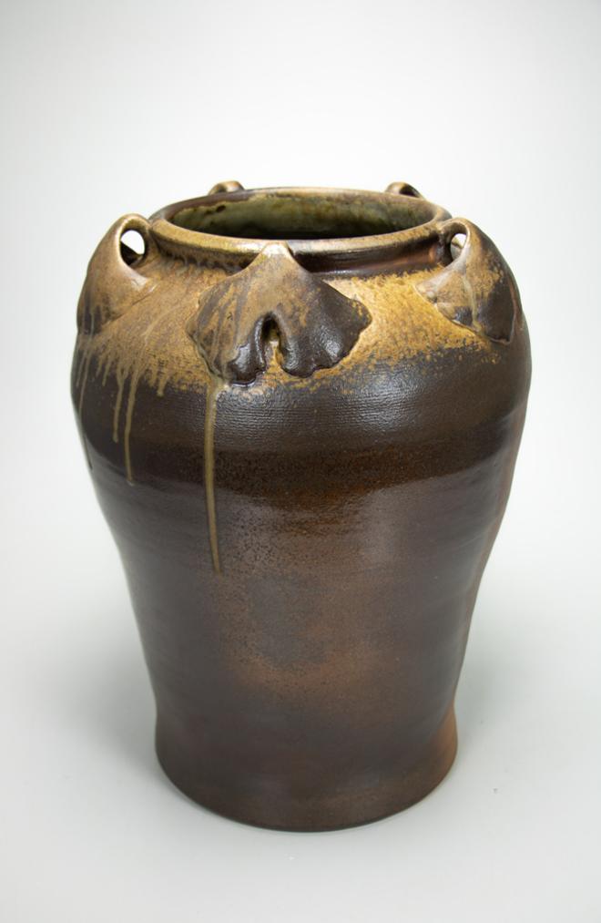 A large, dark brown, wood-fired jar with sculpted ginkgo leaf lugs around the neck.  Brown, natural ash glaze accumulates on the leaves and around the top shoulder of the jar, then drips down the side of the pot.   
