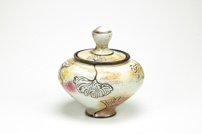 A dark wheel thrown stoneware jar that is covered in white slip with carved ginkgo leaves that wrap their way around it.  Subtle blushes of orange, yellow, tan, and pink can be seen on the white surface.    