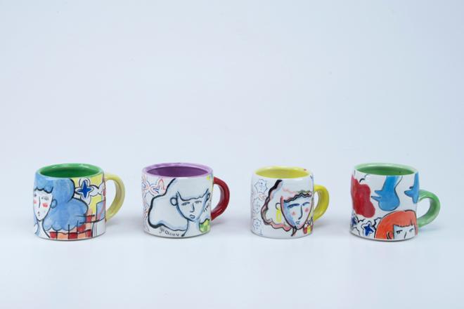 A set of four mugs arranged in a row. Each mug has different drawings on them with a different colored handle and inner glaze.