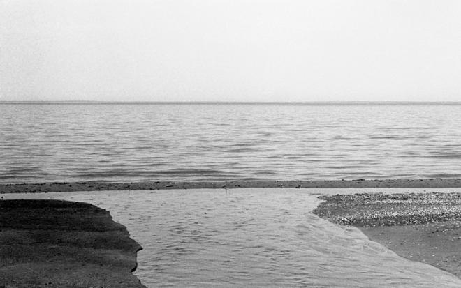 A black-and-white photograph depicting a wide stream of water surrounded by sand on both sides that leads into a larger body of water. The horizon and part of the cloudless sky is visible in the upper half of the photograph.