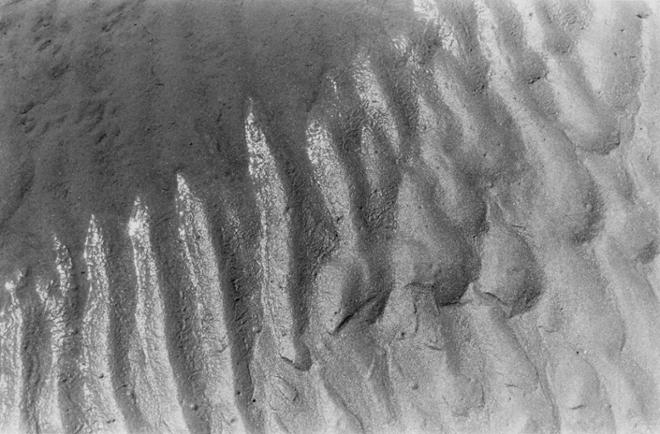 An up-close black-and-white photograph of ridges in wet sand under harsh sunlight. A shadow is present in the upper left-hand corner. 