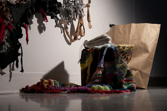 Arranged on a diagonal, starkly lit in a dark corner, is a pile of multiple paper and fiber art pieces. On the wall is a glimpse of a large work made of knotted fibers. From left to right on the floor is a colorful crocheted beaded object draped over an indistinguishable book, with a white, receipt paper book balanced on top. It’s cover is flipped open. Behind the arrangement is a brown paper bag standing upright