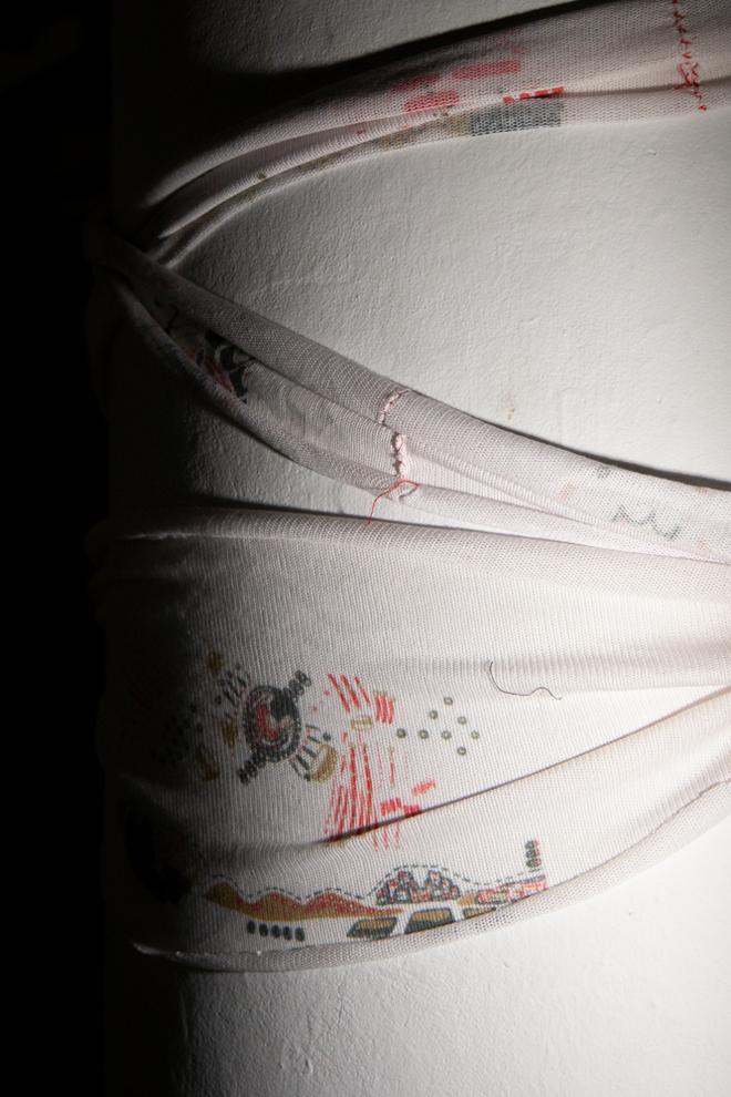 Starkly lit in a dark room, a long strip of white fabric with little threads hanging off is wound around a white pole, allowed to roll and obscure the printed imagery.