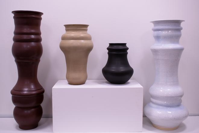 A grouping of four vases in a line on pedestals. The two tales are on opposite ends on the ground and the two smaller vases are on a pedestal in between the tall vases. From left to right, the color of each vase is: Maroon, tan, matte black, and crystalline white.