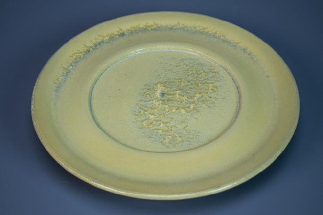 A dinner plate sized plate elevated off the table by 2 inch pedestal foot. The glaze is a matte tan/yellow crystalline glaze with the majority of the crystals grouping in the center of the plate.