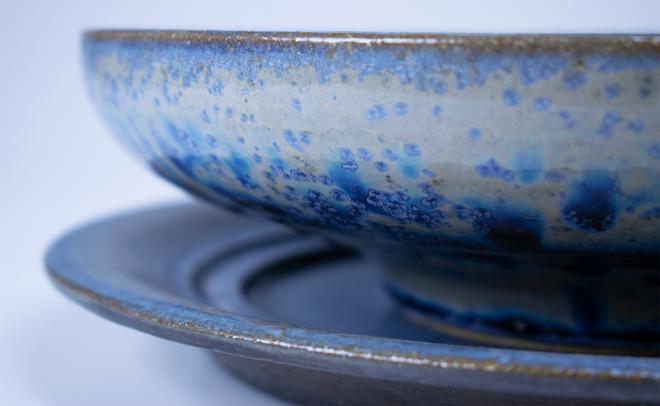 A close up on the blue crystalline glaze of a bowl and plate pair. The image zooms in on the blue crystals on a grey background on the outside of the bowl.