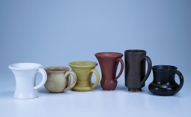 A grouping of different color glazed mugs in a line. There are six mugs and from left to right the colors are: matte white, speckled matte tan, matte yellow, matte maroon, speckled matte dark brown/grey, and semi-matte black.