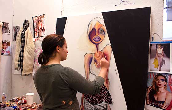 Carrie standing in studio, back to camera, painting large painting of woman