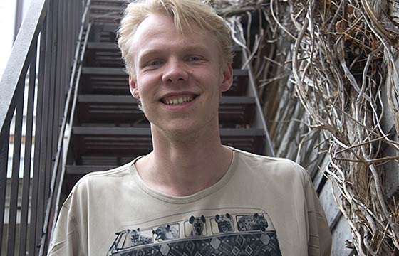Josh standing in front of staircase covered on one side with vines. smiling, spiked blond hair