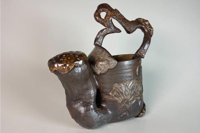Dark ceramic watering can with loopy handle and complimentary carvings surrounding the base of the from. These carvings are brought out by a lighter hued shino glaze.