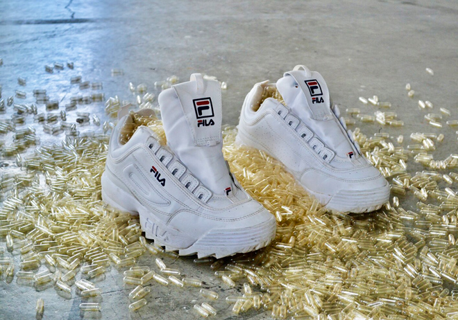 Gold tinted empty pill capsules fill white Fila brand sneakers and spill onto the floor around them.
