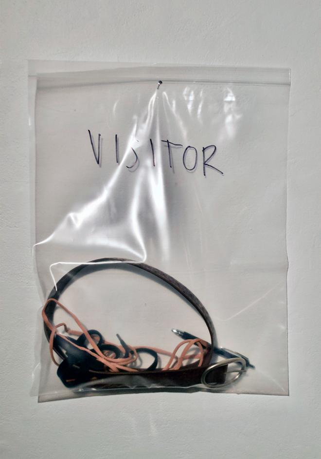 Detail of one of two resealable plastic bags labeled “VISITOR”. The bag is nailed to the wall, and filled with a belt, shoelaces, a sweatshirt drawstring, and a pen. 