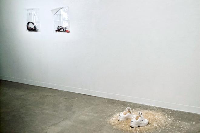  Installation view shows two resealable plastic bags labeled “VISITOR” are nailed to the wall. They contain headphones, belts, shoe laces, sweatshirt drawstrings, keys, wallets, and a lighter. The third resealable bag holds dirtied white shoelaces taken from the Fila sneakers drowning in empty pills on the floor. 