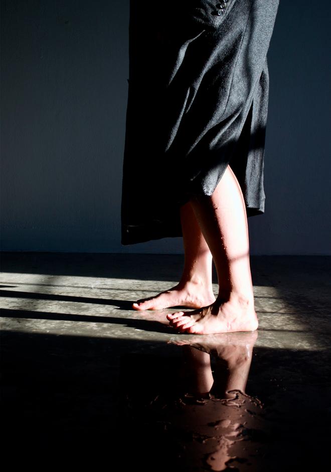 Performer wears soaking wet wool coat. Water drips down her legs and forms a puddle at her feet.