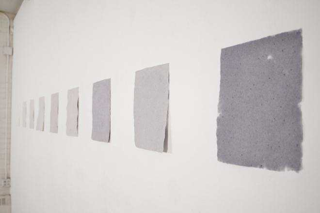 Detail shot of handmade paper hanging on wall. These nine letter sized pages range from blank white, to dark gray creating a grayscale. 