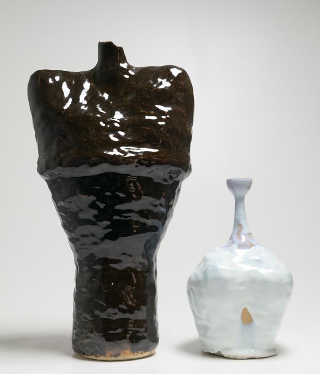A tall vessel that has a cylinder bottom portion, then flattens out and widens into a shape resembling some shoulders. Around the midsection of where the ribs would be there is a bump around the shape meant to resemble breath coming in covered in a dark brown almost black glaze.