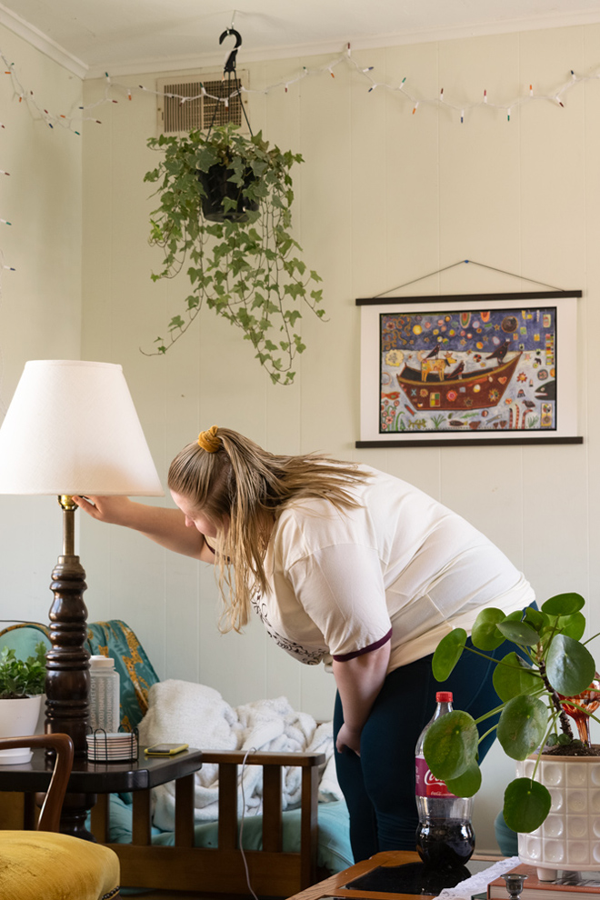  In the center of the photo a woman bends down to turn off the lamp in the living room, she has a white shirt one, a yellow hair tie, and the background has a painting on the wall. 