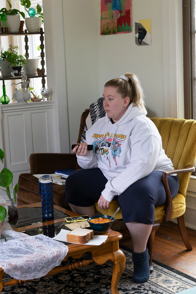 In the center is a girl in a white hoodie, navy pants and navy slipper socks on. In front of her is a bowl of spaghetti O’s, some white bread, her glasses and phone. In her hand is the remote for the TV. She lounges in an old looking mustard velvet chair. 