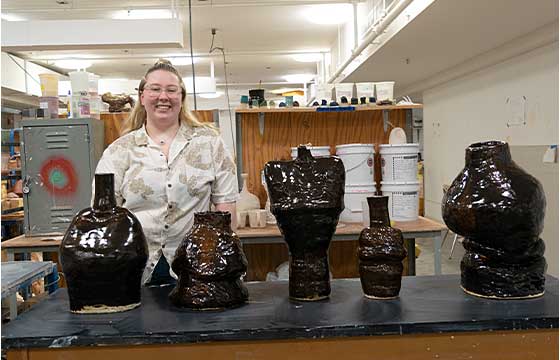 Danielle standing in white sweater behind table stacked with her pottery