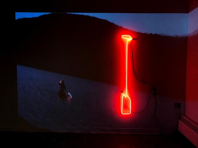Still of a video projected over a red neon paddle install on the wall. The video features a person in a raft, floating on  water, at dusk, using the neon paddle. 