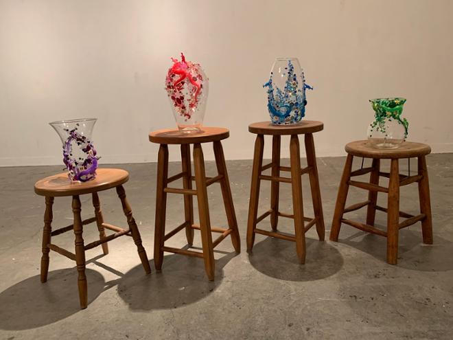 A picture of the same four wooden stools with the vases on them all in a line. In order from left to right the colors are purple, pink, blue, and green.  