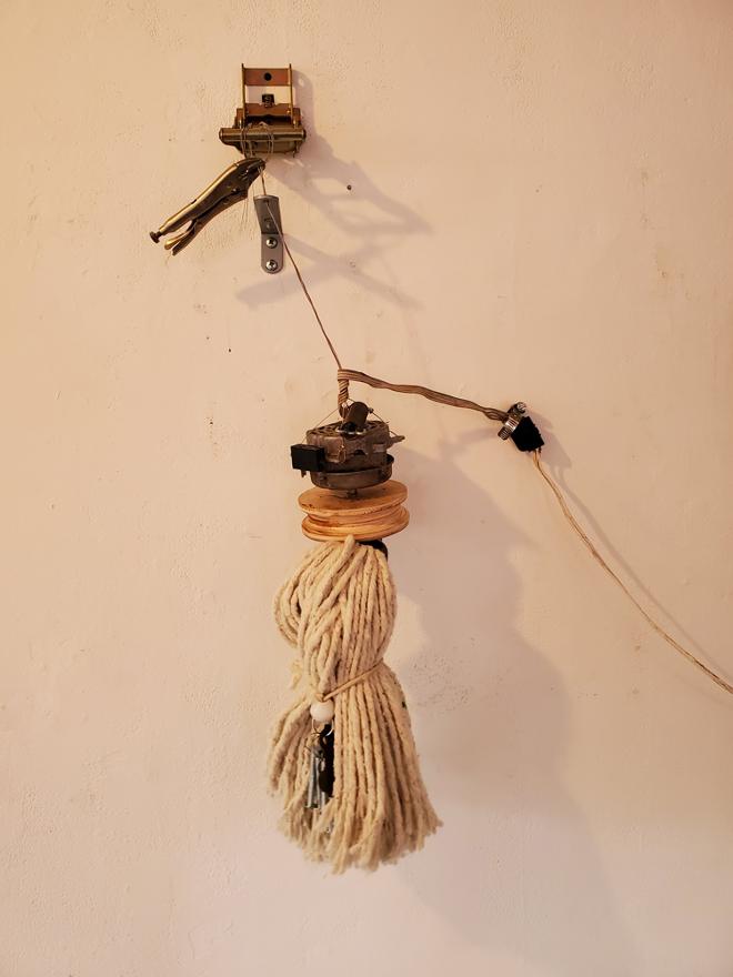 Mop head mounted on a piece of wood attached to a box fan motor, held in place by guitar strings mounted to the wall bracket and ratcheting mechanism, held by vice grips. Electric wire runs off screen.  