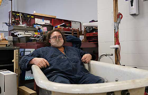 June sitting in cast iron bathtub wearing coveralls and lace mask