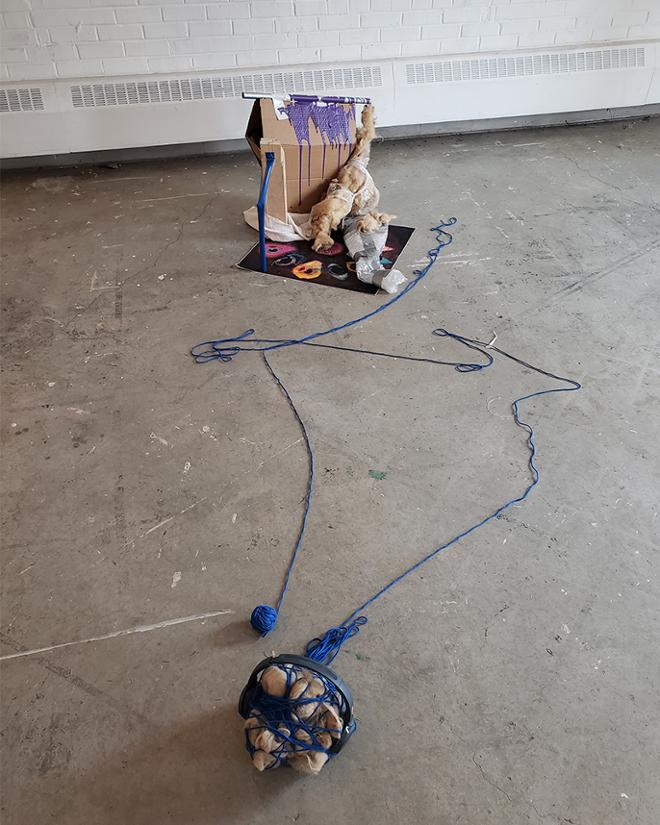 A ball of sheeps wool is tied up with yarn wearing headphones about 8 feet in front of a reclining torso with a wool arm and seran wrap legs, it is wearing a loincloth. 
