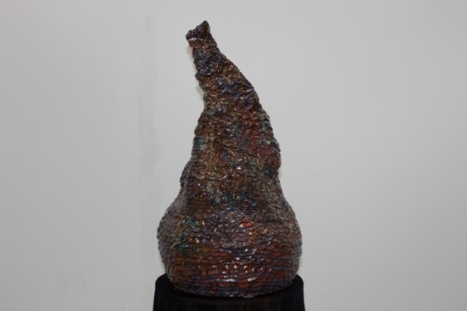 Small coil built vessel that looks as though the top has been squeezed like a water bottle. The texture of the piece itself is made through the pressing of coils. The work itself stands on a table covered with stretchy black fabric. The glazes used are earthy shades of glossy brown, red and some yellow with hints of blue and orange. The sound is broken up to play intermittently within the piece. As for the tone, it leans more toward ambient noises with a mix of gathered music and voice. 