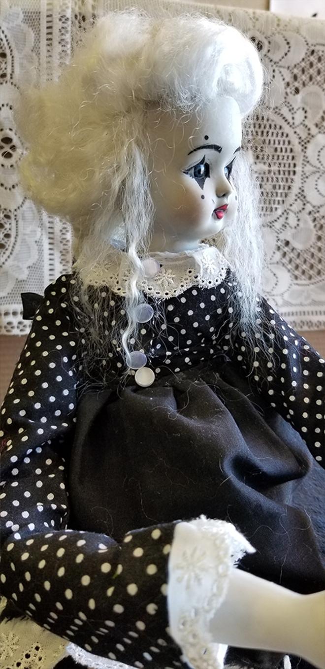 A close up of the face of a porcelain doll sculpture with extended limbs, handmade clothes, and a painted face.  