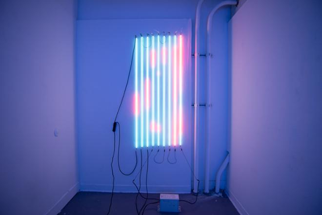 Nine clear tubes pumped neon mercury each five feet tall. Shifting from red to a light blue light. Hung vertically on a white wall.  Each tube is spaced three inches apart from each other across a span of two feet. 