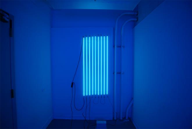 Nine clear tubes pumped neon mercury, hung vertically on a white wall.  Each tube is spaced three inches apart from each other across a span of two feet horizontally.  The light from the tubes is filling the entire space in a light blue light. 