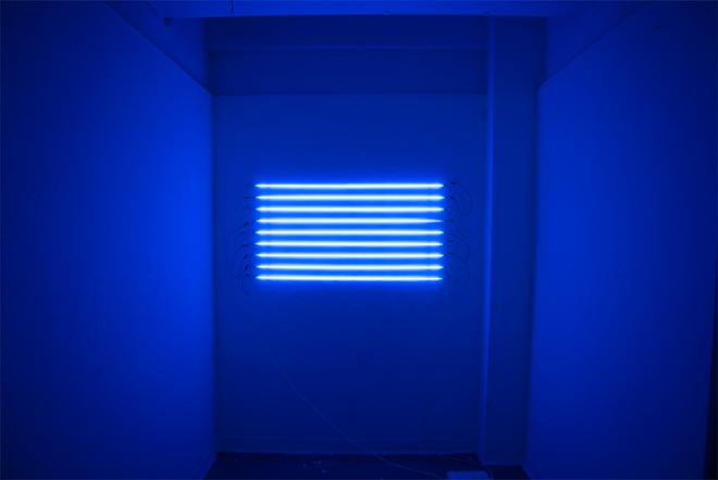 Nine blue tubes pumped argon mercury, hung horizontally on a white wall.  Each tube is spaced three inches apart from each other across a span of two feet vertically.  The light from the tubes is filling the entire space in a blue light. 