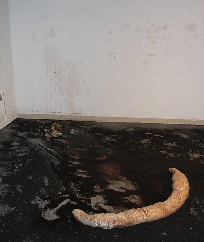 Black glass abrasive is on the ground, a lumpy rolled form made of wood shavings wrapped in plastic sheeting about 4.5ft is curved in a slight crescent shape on the ground pushed towards the center of the gallery space away from the splash on the wall.  There is a red watery liquid on the ground and splattered on the wall as high as 6 feet.  Below the splatter there are about 8 smaller rock pieces and dents in the gallery wall above and around the splatter.