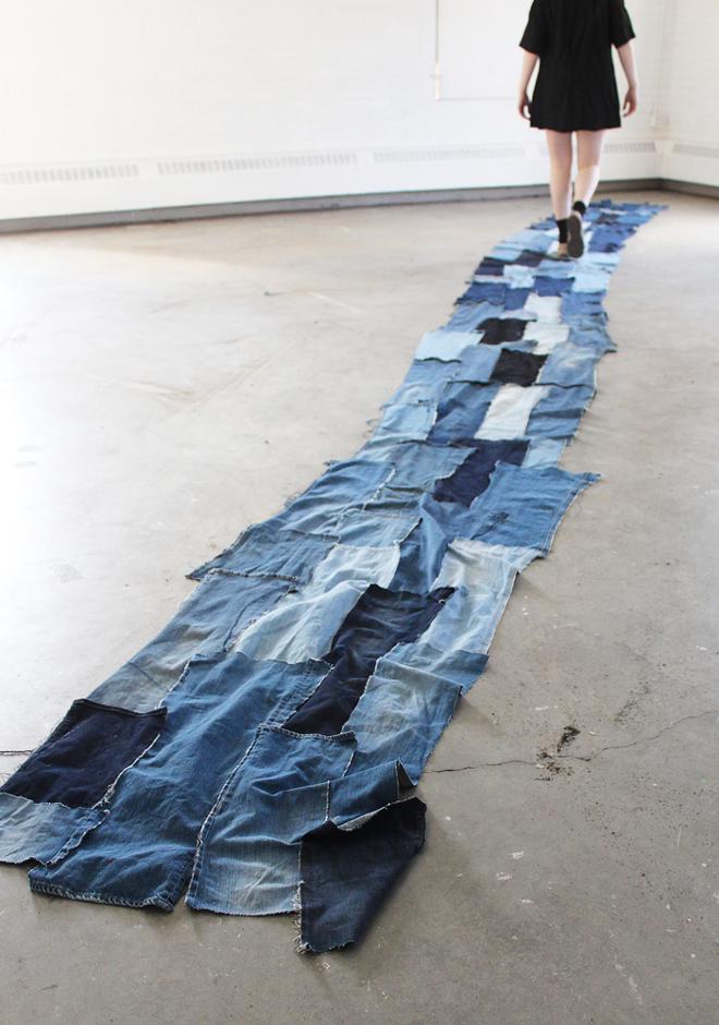 A very long runway created entirely of thread and recycled jean pant legs with a figure at the end dressed in a black dress and black socks walking a stereotypical runway model walk.