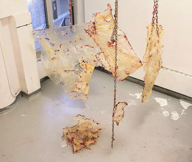 3 large sheets of naturally cured gelatin and 1 smaller sheet hang from chains in front of a window with a pile of gelatin skins on the ground below it all adorned with the red coloring made up of silicone and red iron oxide to enhance the holes in the sheets that look like cuts and the chain is rubbed with that mixture as well.