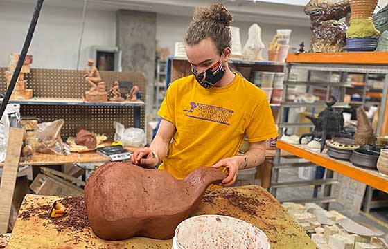 Vincent in studio wearing yellow AU Simming shirt, carving large terra cotta sculpture resting on a piece of foam