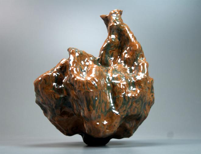 A small, glossy brown and turquoise vessel that balances on a narrow definite point while exhibiting a lumpular brain-like body and a neck that reaches up and off to one side.