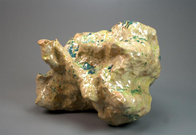 A lumpy vessel of stony color lies on its side and exhibits a surface adorned with glossy light chartreuse and spots of deeper greens and blues. Like a jiggling green Dalmatian if it was a pot.