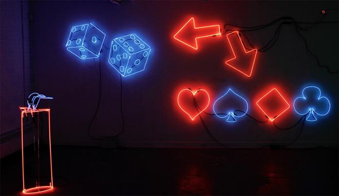 A full wall installation consisting of a neon sign in the bottom right corner that has the 4 card suits alternating in color between red and blue. In the top left corner there is blue neon dice. Placed between these two signs are orange neon arrows pointing at both signs. In the foreground is a neon lighter sculpture.